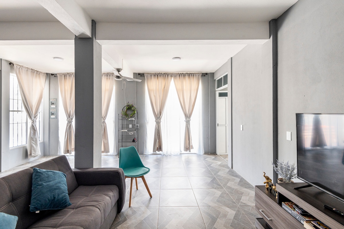 The living room features large windows draped with soft, neutral-toned curtains that allow natural light to fill the space. A comfortable sofa with a decorative pillow is positioned opposite a wall-mounted television. A small accent chair adds color and style, enhancing the room's inviting design.