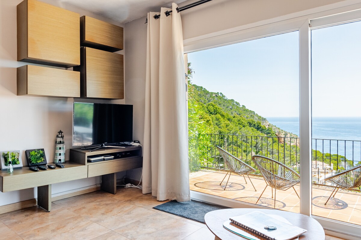 A spacious living area is highlighted by a large glass door offering views of the sea and surrounding landscape. A television is mounted on a contemporary media console, while cozy seating is provided by woven chairs. Natural light fills the space, complemented by soft curtains.