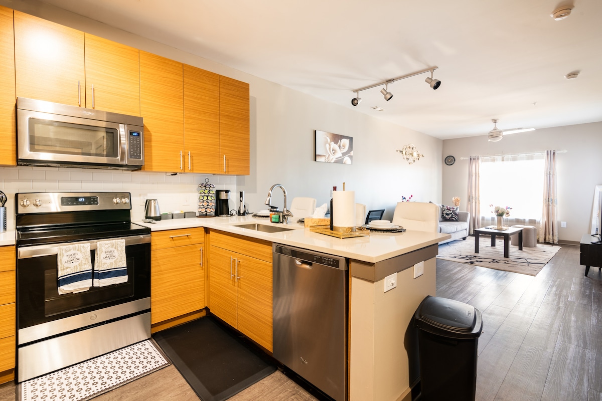 The modern kitchen features wooden cabinetry and stainless steel appliances, including a microwave and dishwasher. Counter space is available for meal preparation, with a small dish rack present. The open layout extends to a living area, enhanced by natural light from a nearby window.