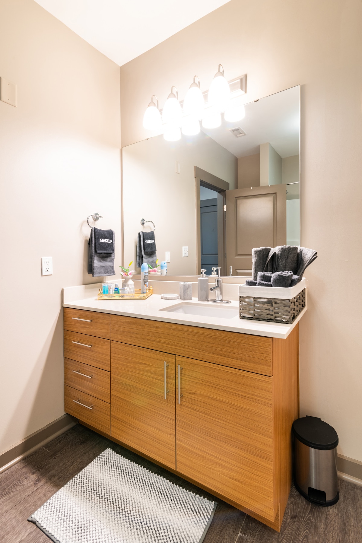 A well-appointed bathroom is displayed, featuring a modern vanity with a wooden finish. A large mirror is positioned above the sink, complemented by ambient lighting. Essential toiletries are neatly arranged on the counter, and a small basket holds towels. A textured mat is placed on the floor.