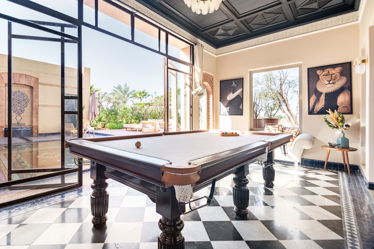 A spacious room designed for leisure features a classic billiard table, surrounded by elegant furnishings. Large glass doors provide a seamless transition to the outdoor area, allowing natural light and views of the garden to enhance the atmosphere.