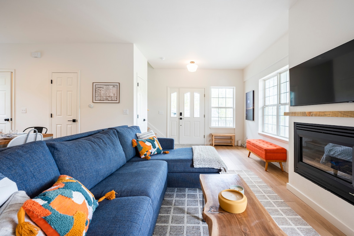 A spacious living area features a large blue sectional sofa adorned with colorful throw pillows. A sleek television is mounted on the wall above a modern fireplace. Natural light floods in through large windows, highlighting the light wood flooring and simple decor.