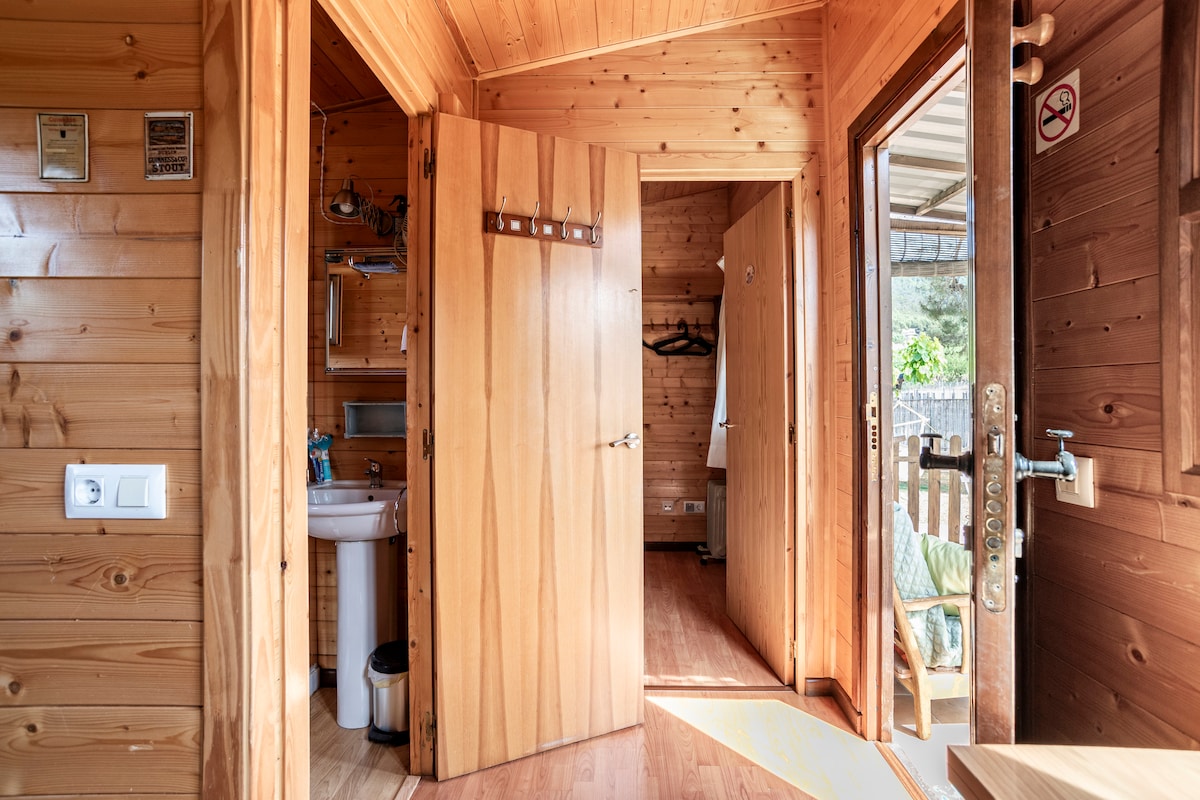 A wooden interior features natural wood paneling, with doors leading to separate areas. One direction provides access to a bathroom with a sink, while another door opens to an outdoor area. Hooks for hanging items are mounted on the wall.