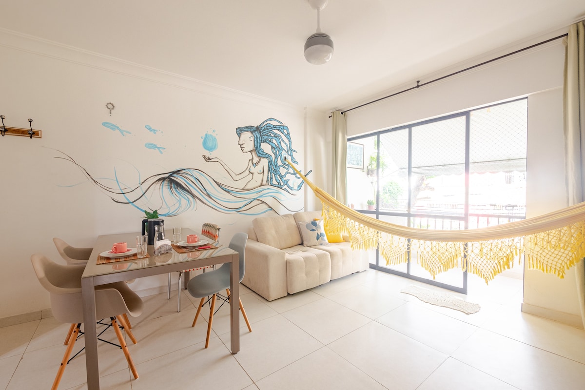 The living space includes a dining table set for two with soft, neutral tones and a cozy sofa. A colorful mural of a figure and aquatic elements adorns the wall. A hammock is draped beside a large window, allowing natural light to fill the area.