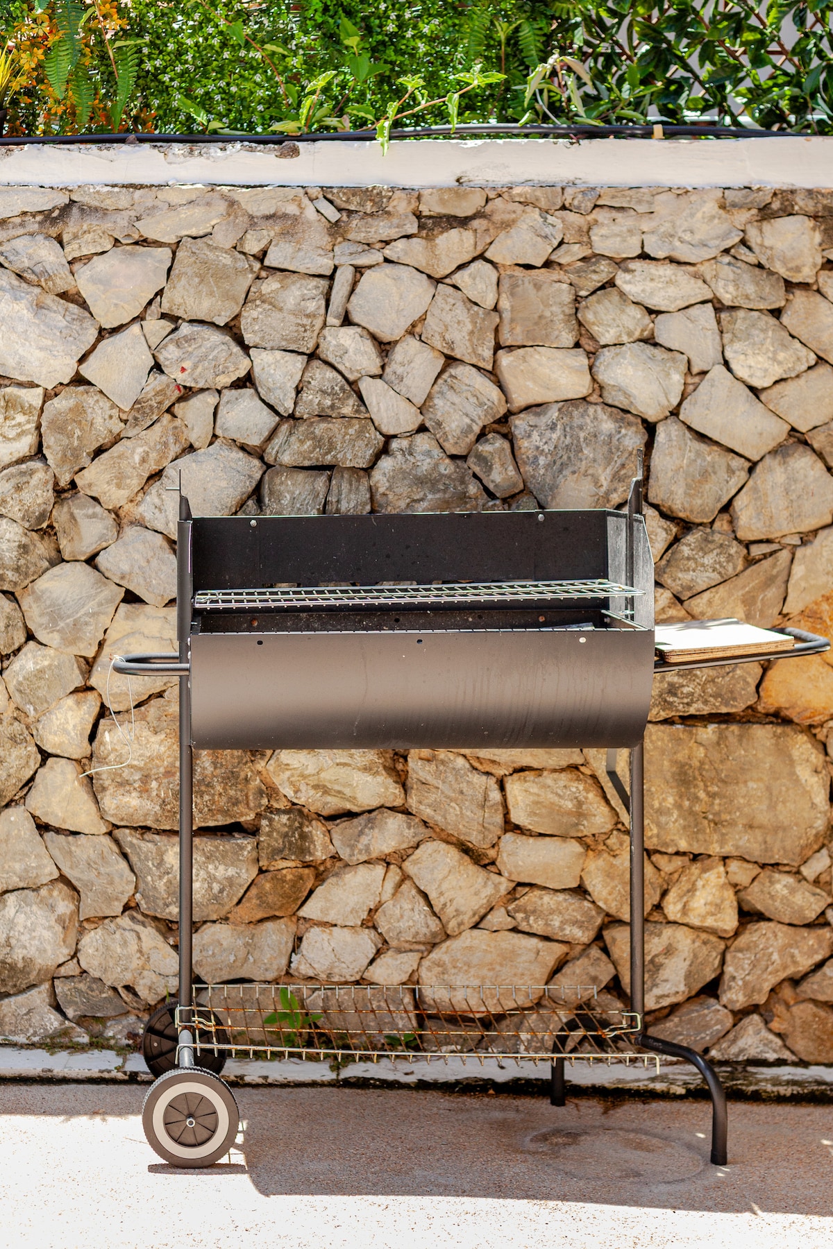 A charcoal BBQ grill is positioned against a natural stone wall. The grill features a cart with wheels for mobility, a cooking surface, and a side table for food preparation.