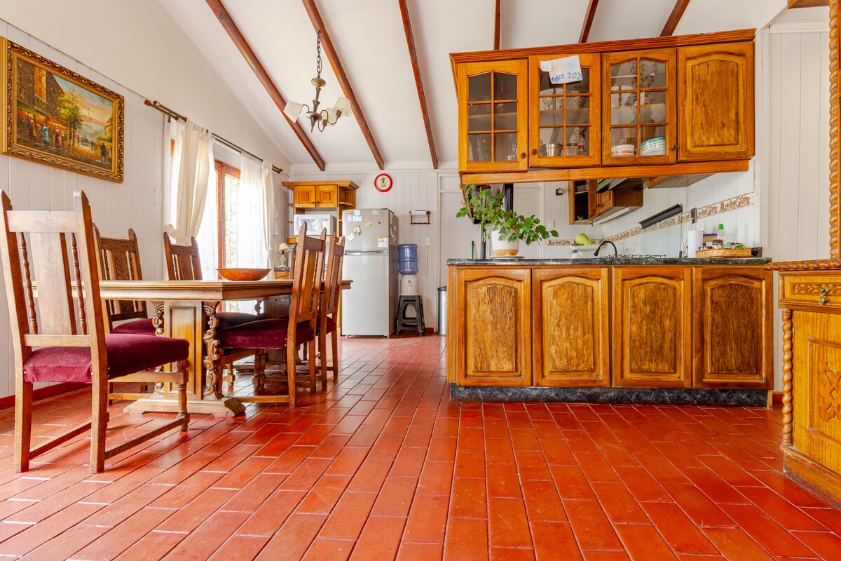 A dining area features a wooden table surrounded by six matching chairs. Light enters through large windows, illuminating the terracotta tiled floor. A wooden kitchen counter is visible in the background, complemented by cabinetry with glass doors. An organized space conveys functionality and warmth.