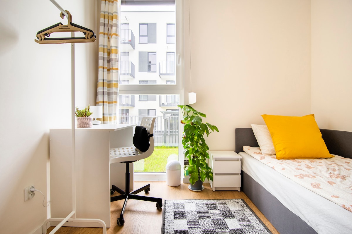 A single room features a comfortable bed with a yellow accent pillow, a modern desk with a chair, and a small plant. Natural light enters through a window overlooking the garden. A rug adds warmth to the wood floor, while a small nightstand completes the space.