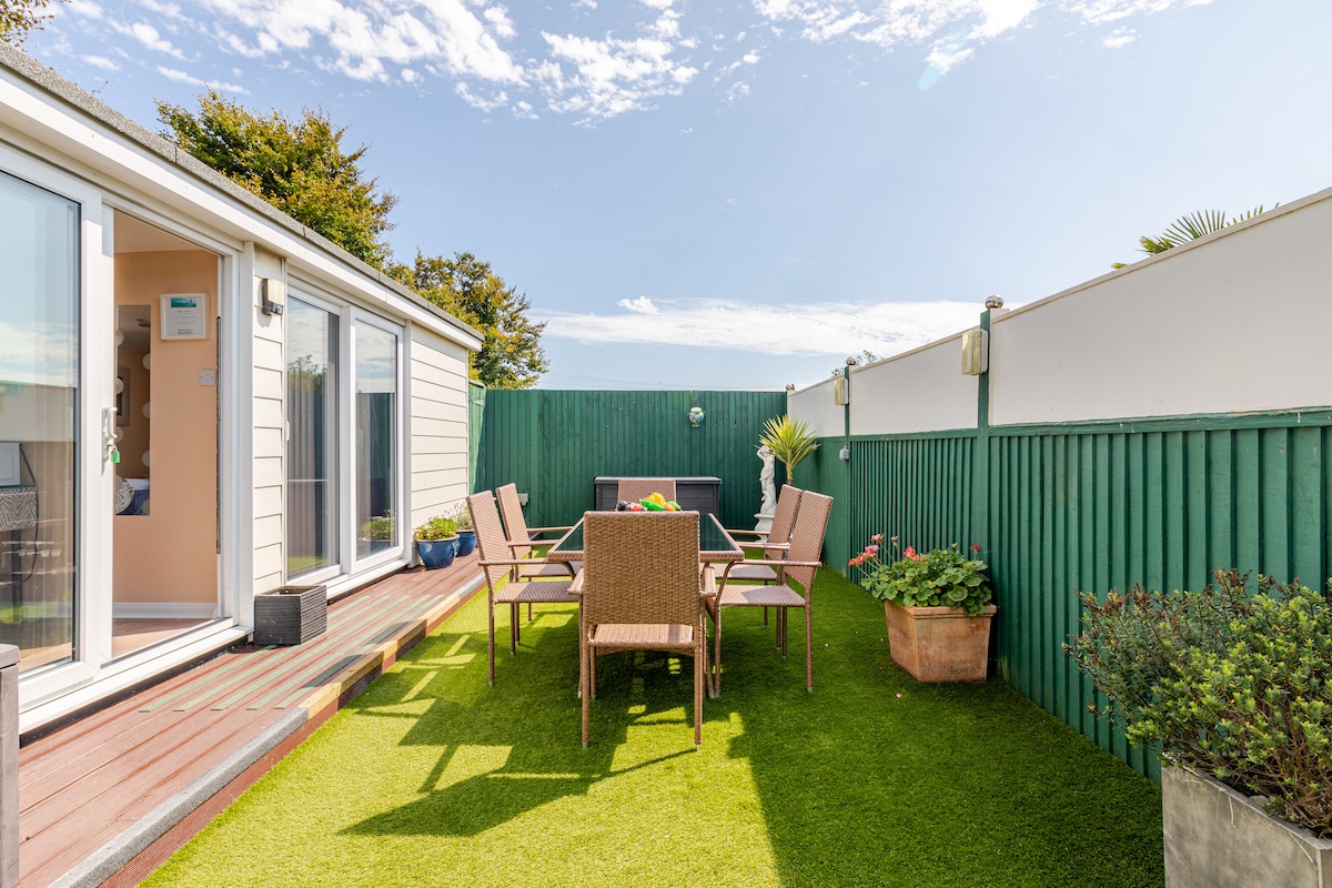 A private decking area is surrounded by green fencing, featuring a table with six chairs positioned on artificial grass. Potted plants are visible, and sliding doors provide access to the interior. Bright sunlight illuminates the space, creating an inviting outdoor setting.