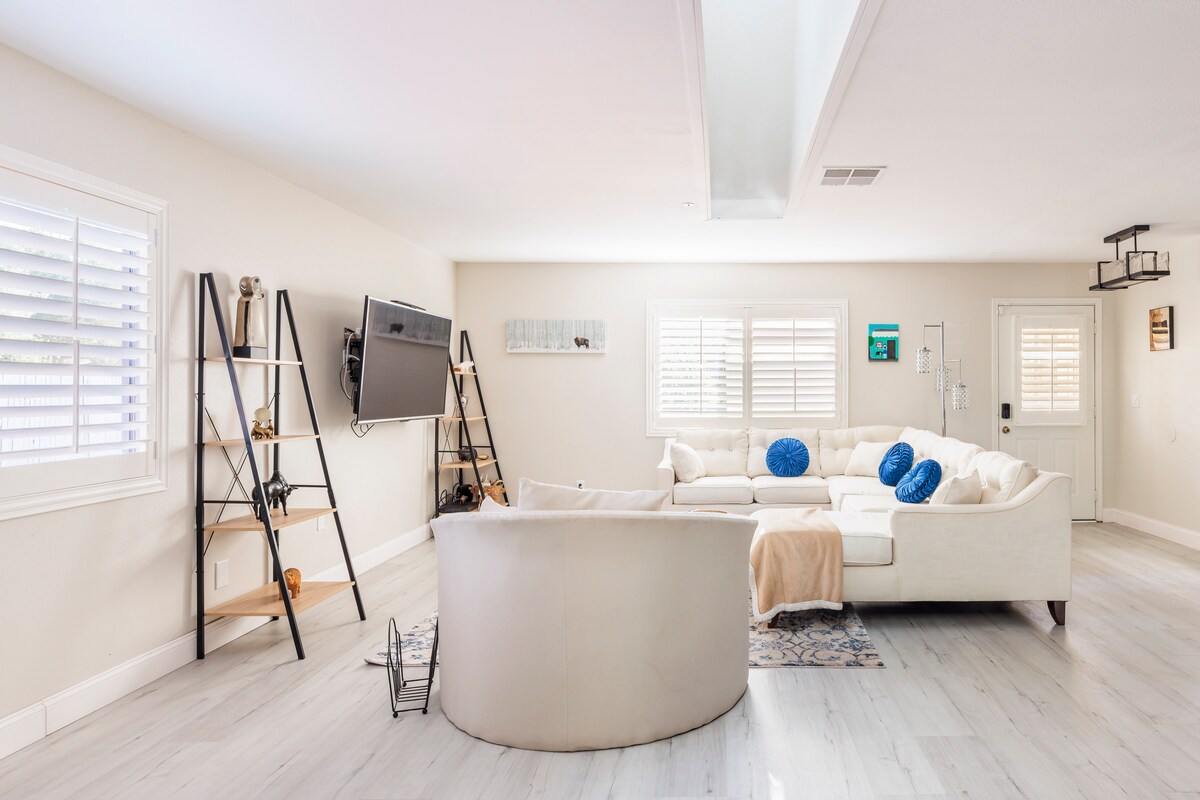 An open living area features a large sectional sofa arranged around a light-colored rug. A television is mounted on the wall, and a ladder shelf displays decorative items. Natural light filters through adjustable blinds, illuminating the space's neutral tones.