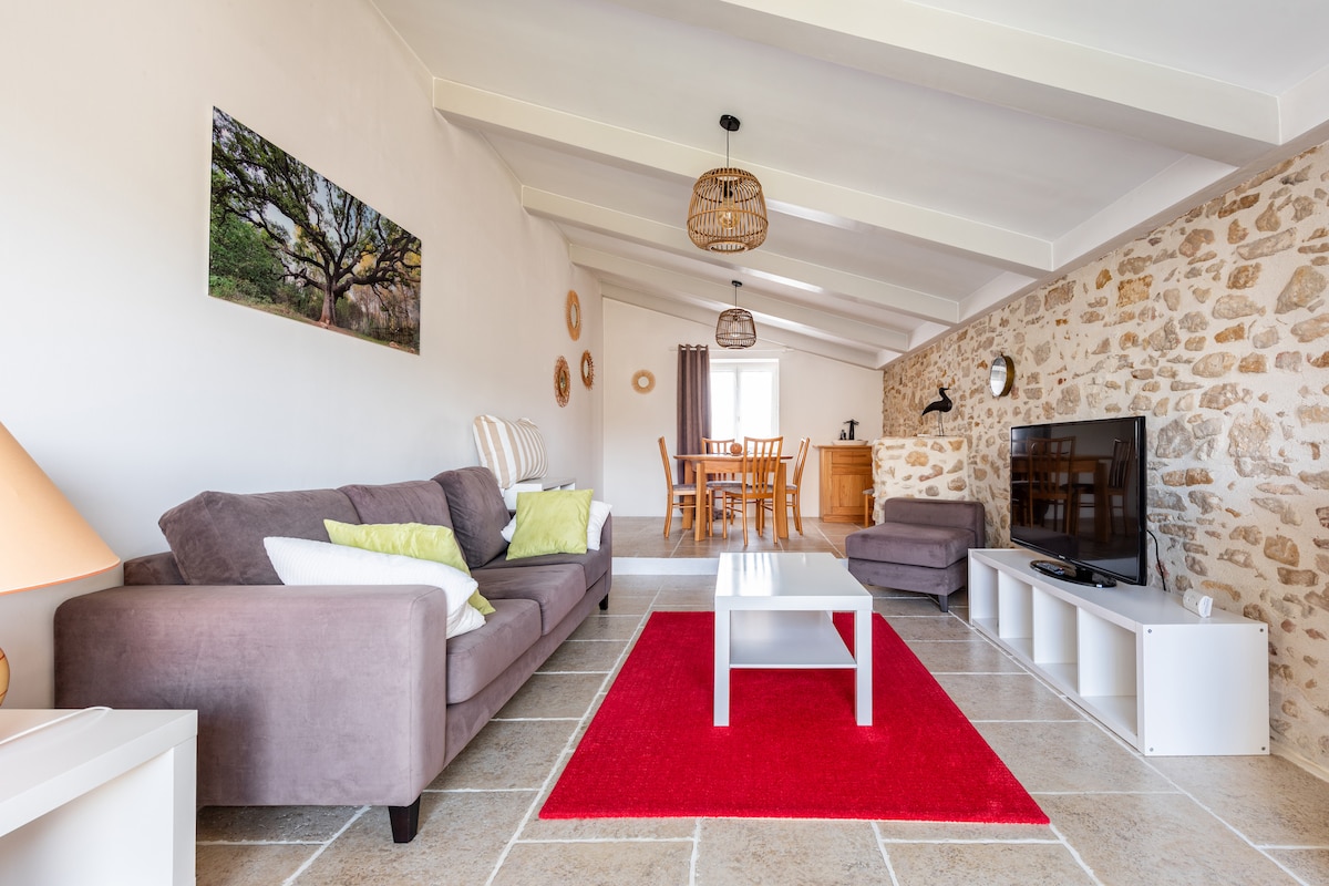 A spacious living area features a comfortable gray couch adorned with green and white pillows, placed on a vibrant red rug. A flat-screen television is positioned against a white media console. A dining area with wooden furniture is visible in the background, complemented by natural light from large windows.