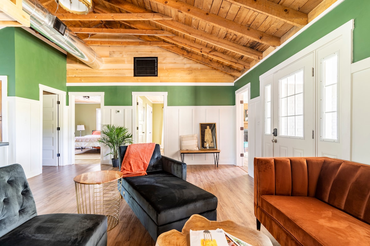 An inviting living area features a mix of seating options, including a dark grey couch and an orange armchair. Exposed wooden beams enhance the ceiling, while vibrant green walls provide a refreshing backdrop. Natural light fills the space through large front doors, leading to additional rooms.