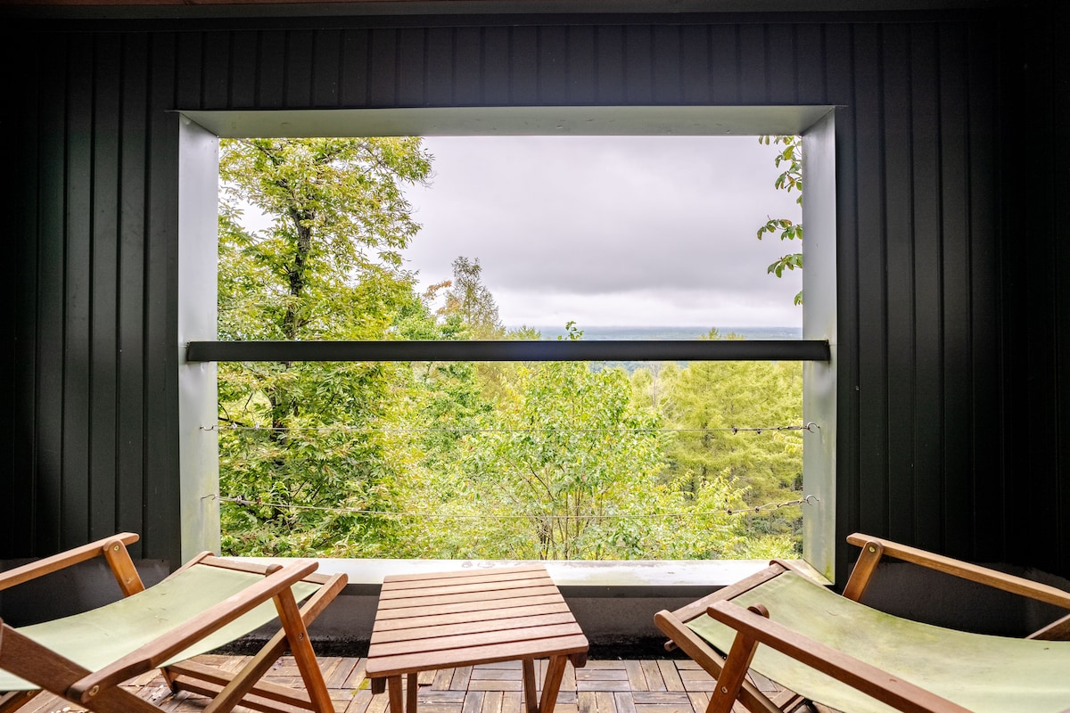 A spacious window frame offers a serene view of the surrounding forest. Two wooden lounge chairs are positioned beside a small wooden table, creating a relaxing outdoor space to enjoy nature.