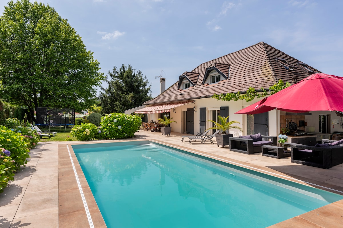 A spacious outdoor area features a clear swimming pool framed by a neat terrace. Comfortable seating arrangements are positioned under vibrant red umbrellas, providing shade. Lush greenery and colorful plants enhance the property's inviting ambiance, under a bright blue sky.