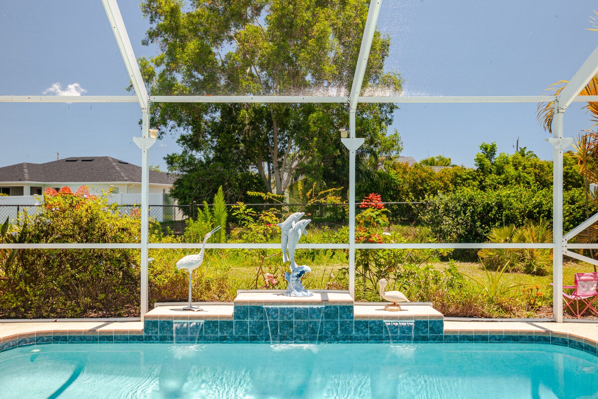 A fenced saltwater pool is framed by a screened enclosure, featuring a decorative statue at the center of the pool deck. Lush greenery and flowering plants are visible in the background, contributing to a serene outdoor environment.