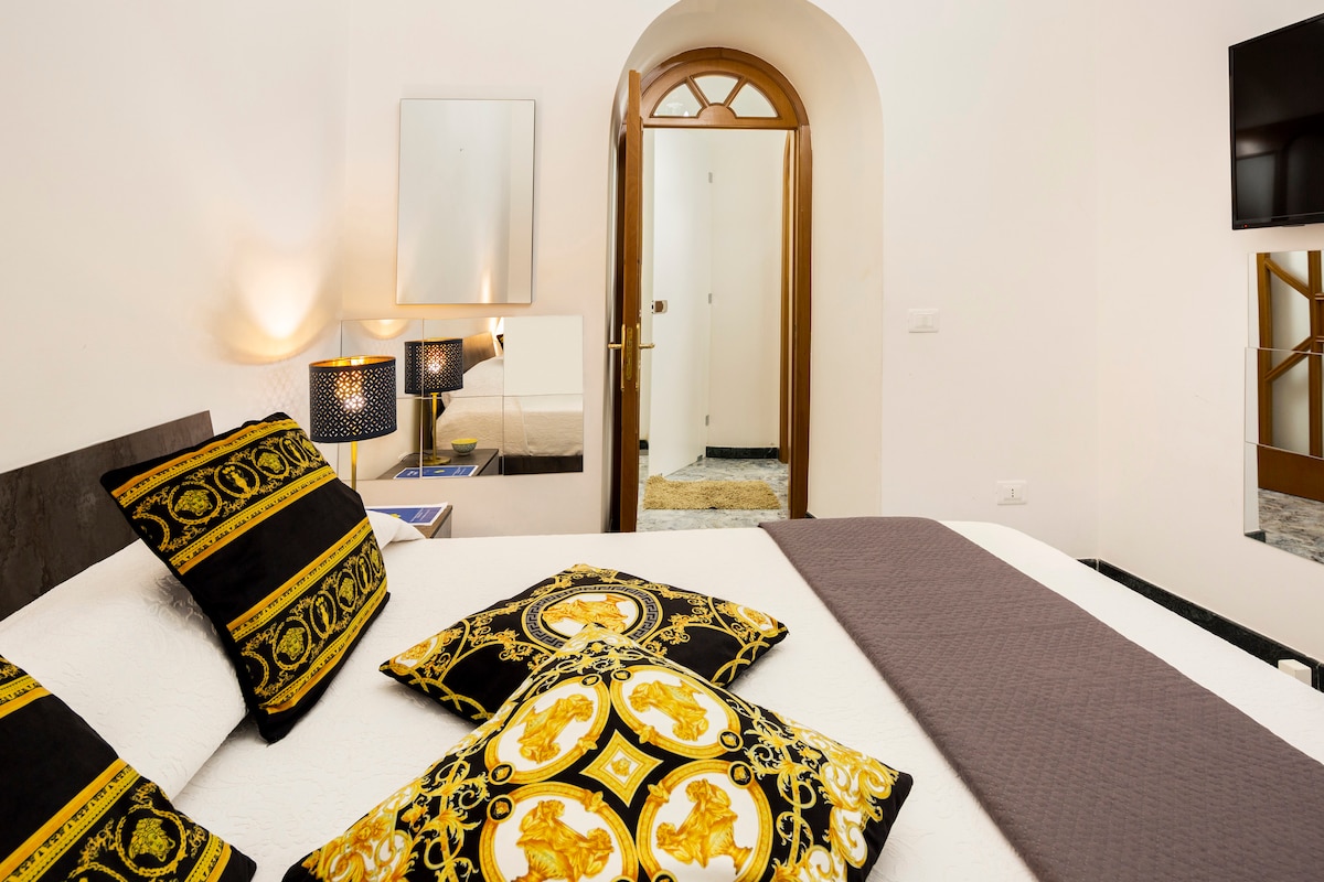 A spacious bedroom features a large bed adorned with decorative cushions in rich black and gold patterns. Soft lighting is provided by lamps on either side of the bed. A mirror and doorway are visible, highlighting the room's layout.