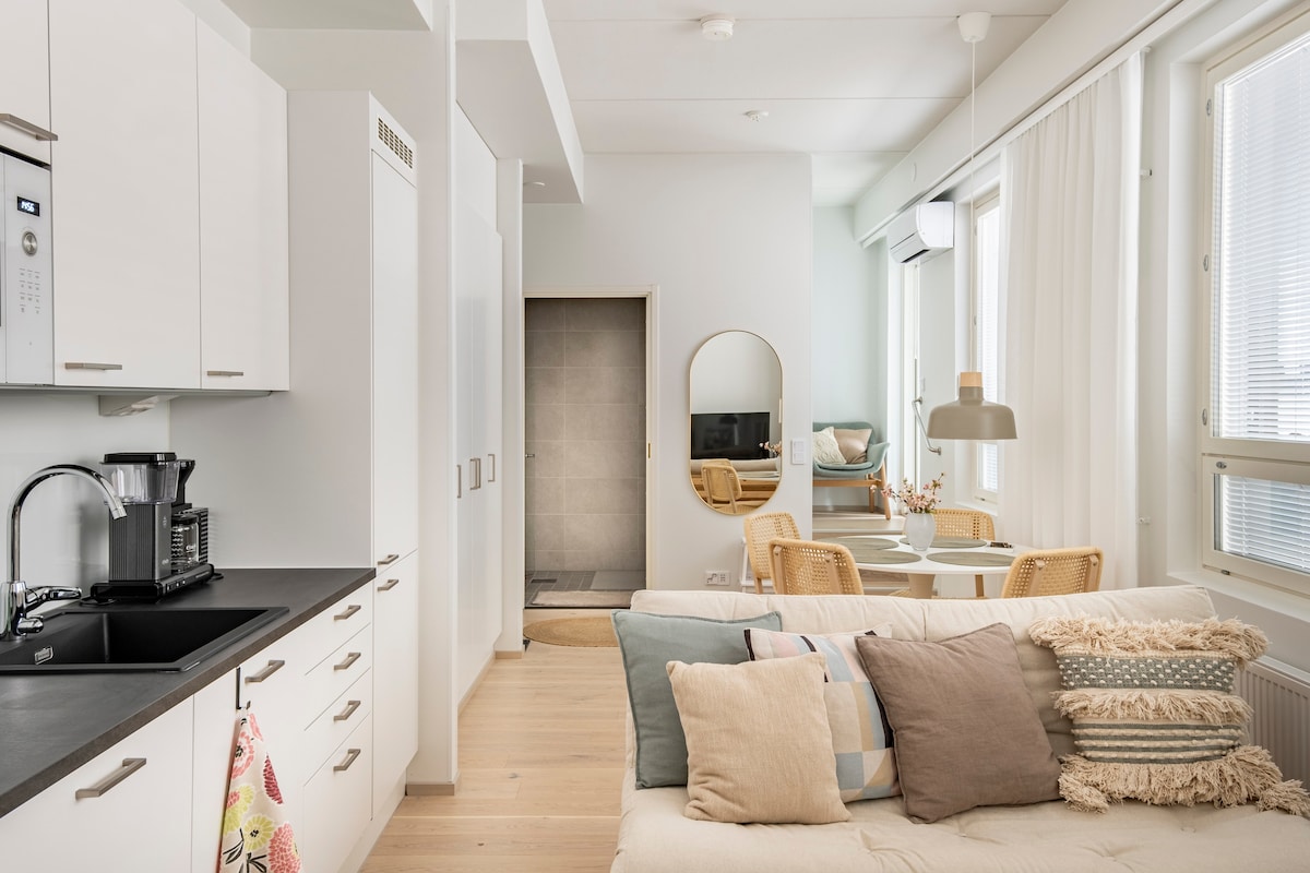 A modern kitchen area is visible, featuring sleek white cabinetry and a dark countertop. A comfortable seating space includes a light-colored sofa adorned with decorative pillows. Natural light floods the room through large windows, with a dining area that accommodates four and a mirror reflecting the relaxing atmosphere.