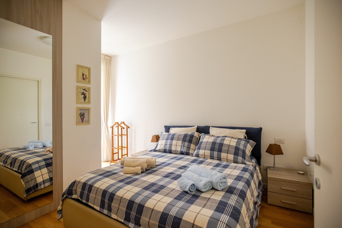 A bright bedroom is featured, highlighted by a spacious bed adorned with a checkered blanket and neatly arranged towels. A bedside lamp sits on a nightstand, and a full-length mirror adds depth. Light filters in through sheer curtains, enhancing the serene ambiance.