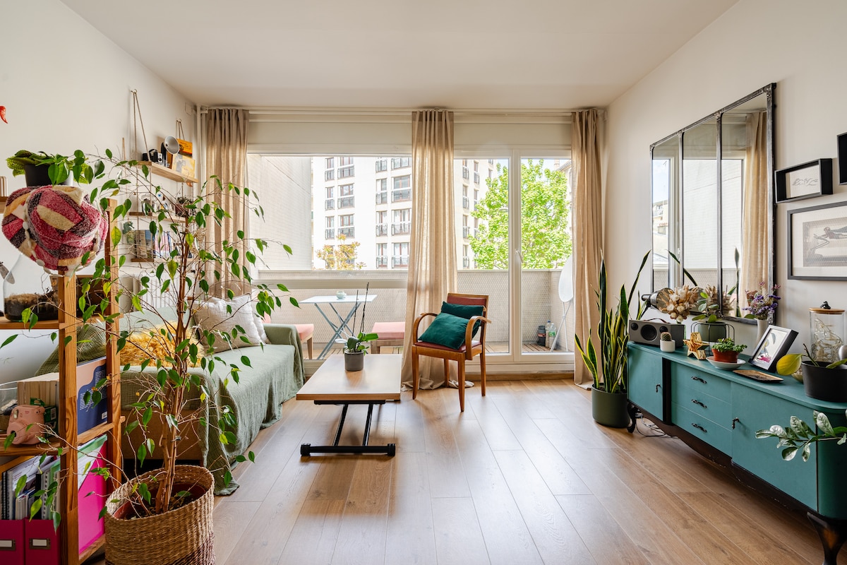 A bright living area is seen with large windows allowing natural light to fill the space. A comfortable chair and a wood coffee table are arranged on light wooden flooring. Green plants add a touch of nature, complementing the cozy ambiance.