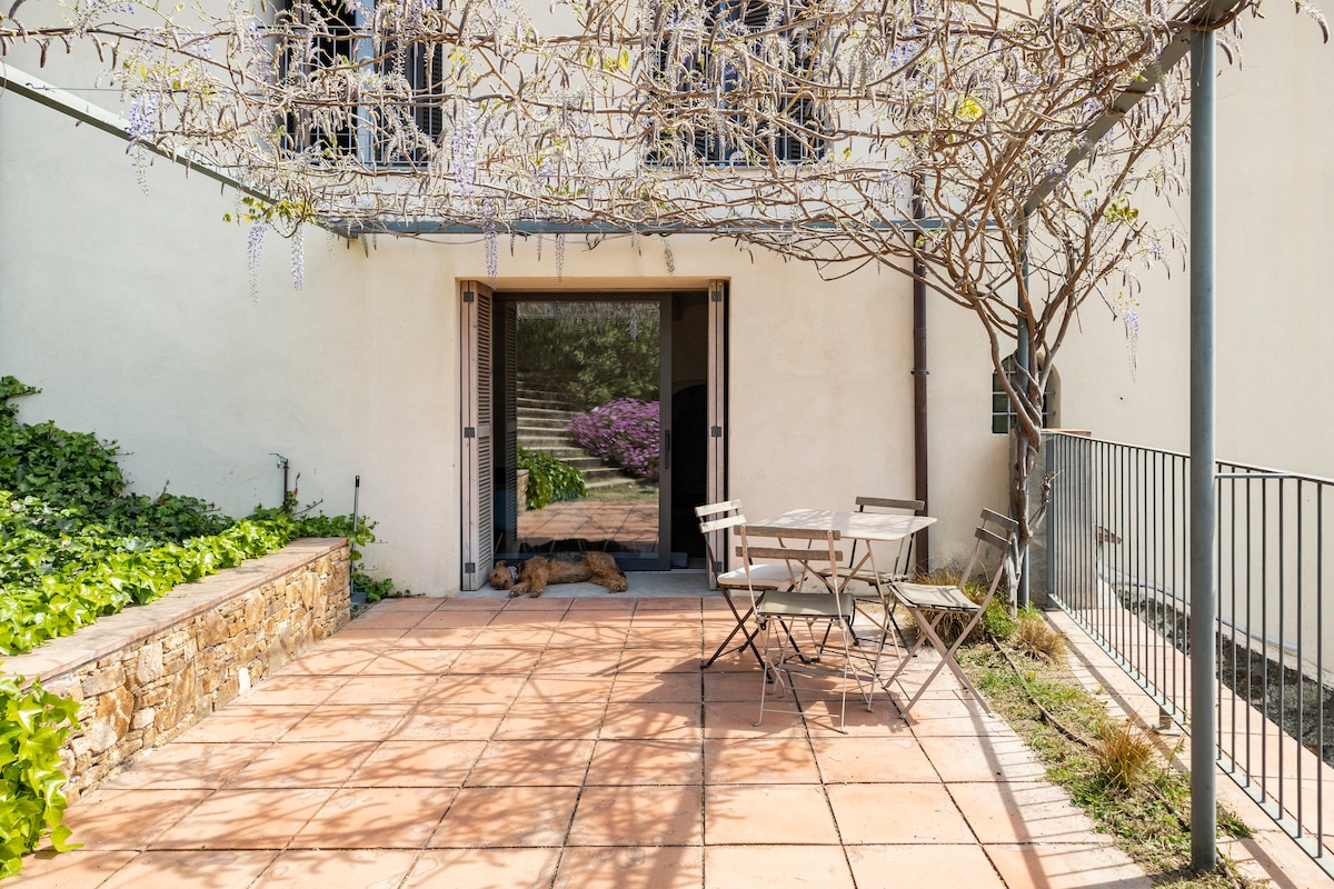 A private terrace features a stone wall and a tiled floor, complemented by a small table and chairs for outdoor seating. Sliding glass doors provide access to the interior space, while shadows of plants and vines create a natural ambiance.