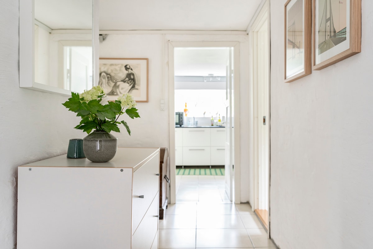 A hallway leads to a kitchen, showcasing white walls and tiled flooring. A console table with a small vase of flowers is positioned against the wall. Framed artwork is displayed on the adjacent wall, creating a welcoming entrance.