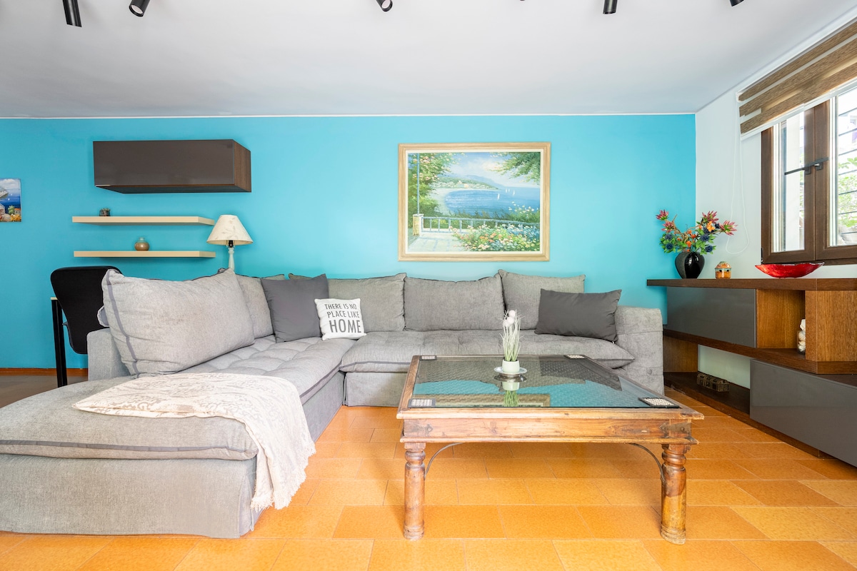 A spacious living area features a large, L-shaped gray sofa complemented by a wooden coffee table with a glass top. A colorful artwork hangs on a vibrant blue wall, and natural light enters through nearby windows adorned with light blinds.