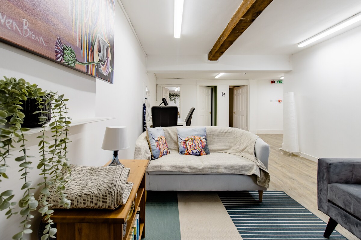 A spacious lounge area features a comfortable sofa adorned with decorative cushions, a wooden side table, and greenery. An artwork hangs on the wall, complemented by soft lighting and a blend of rugs underfoot. A small workspace is positioned towards the back.