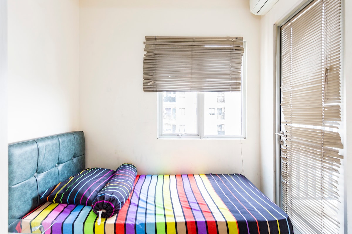 A bright bedroom features a spacious bed dressed in a vibrant striped blanket. Natural light filters in through a window covered with horizontal blinds, providing a warm ambiance. A door leads outside, enhancing the room's accessibility and airy feel.