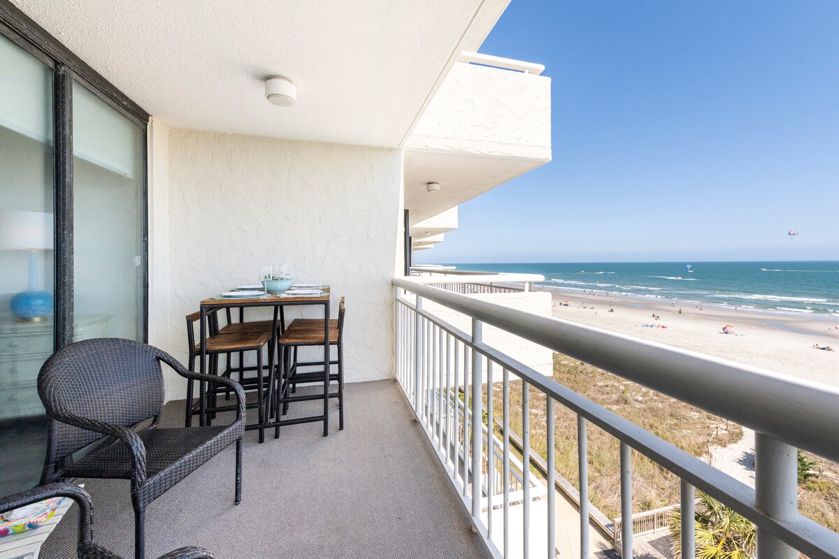 A balcony overlooks the ocean with a clear blue sky. A small dining table with two high chairs is positioned beside a comfortable chair. The sandy beach is visible below, with small figures enjoying the sun and waves.