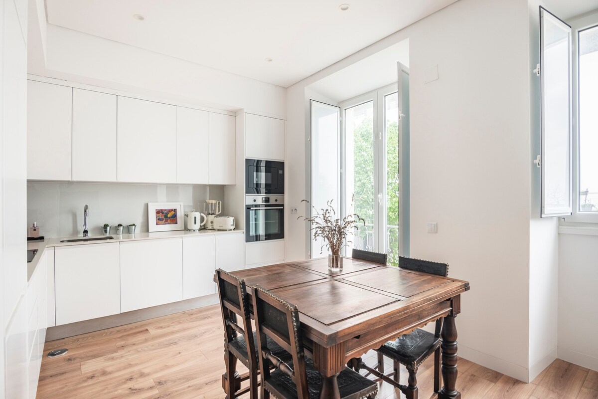 A bright kitchen area features a rustic wooden dining table surrounded by black chairs. White cabinetry blends with modern appliances, and two large windows allow ample natural light while providing views of greenery outside.