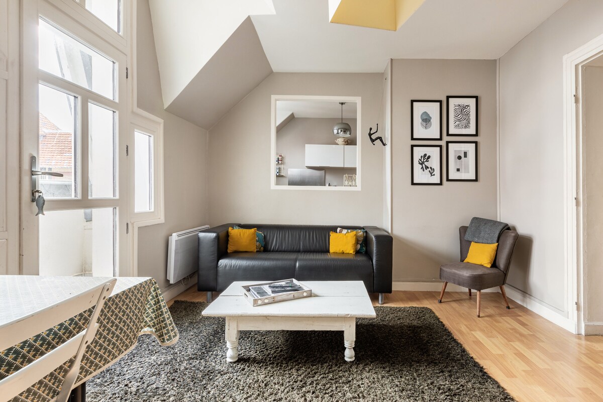 A bright living area features large windows allowing ample natural light. A black leather sofa with yellow accent pillows is positioned next to a light wooden coffee table on a soft, dark rug. Art pieces are displayed on the wall, and a small dining table is visible.