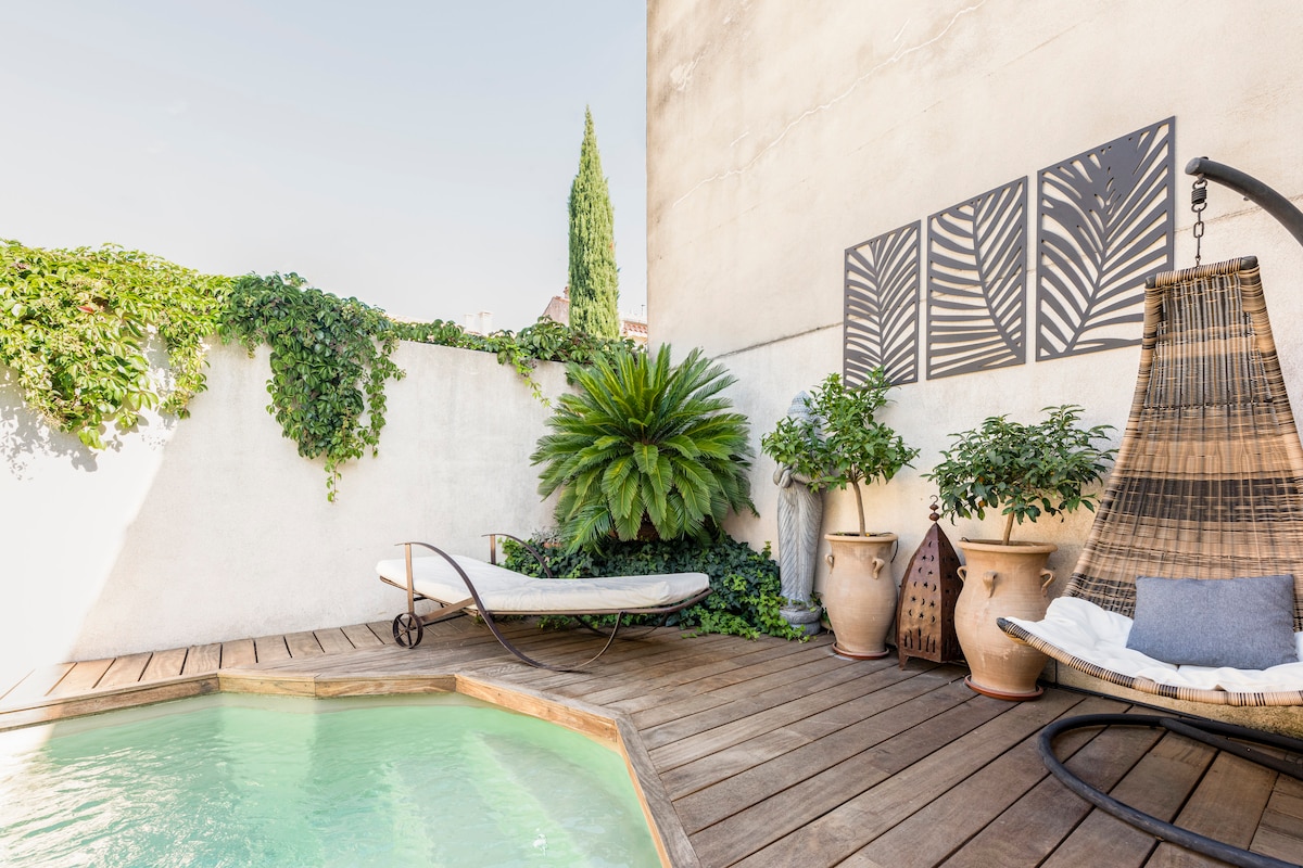 A private wooden deck surrounds a small turquoise pool, accented by lush greenery and a tall potted plant. A hanging chair invites relaxation, complemented by two decorative pots with plants. The wall features a minimalist art piece, enhancing the tranquil outdoor space.