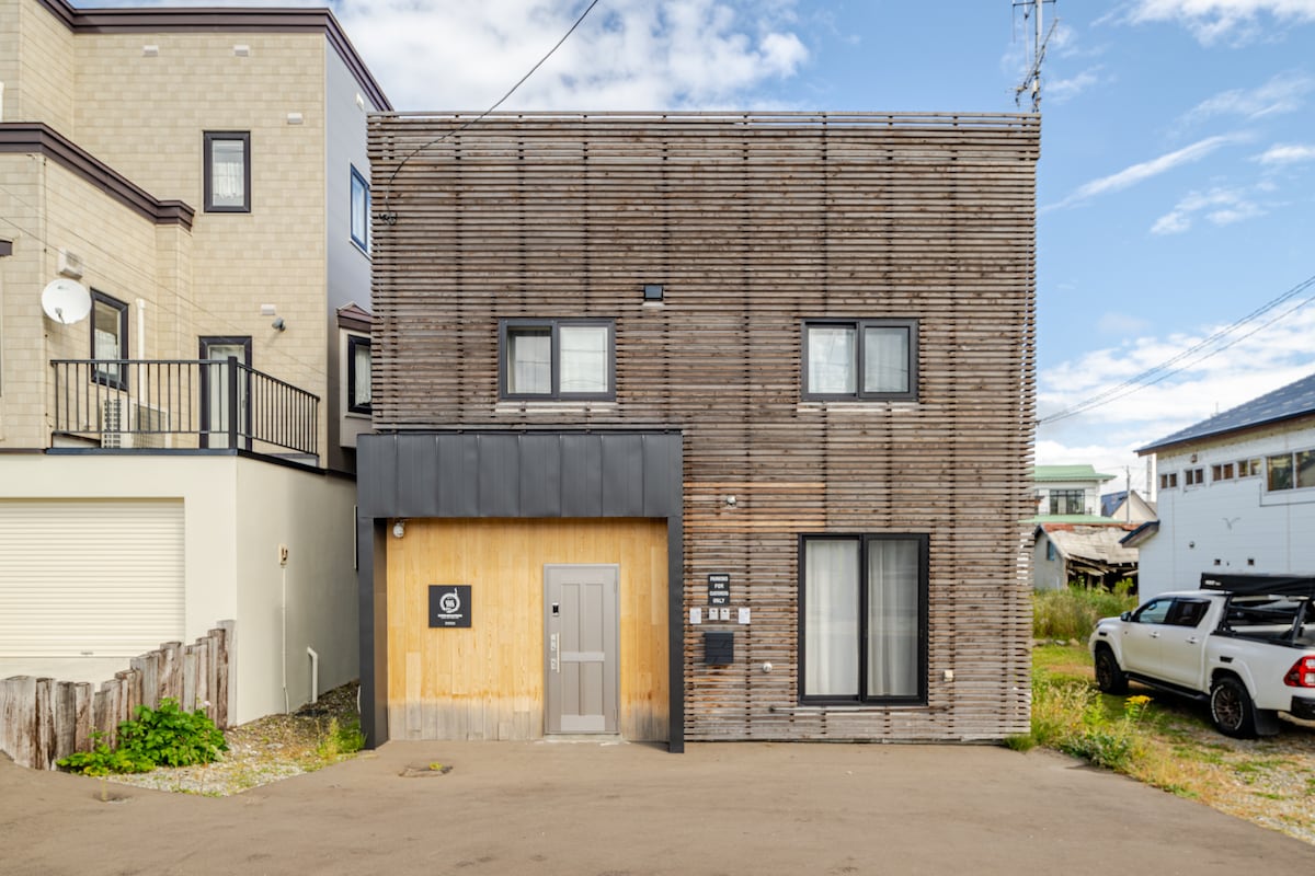 The exterior of a modern villa is presented, featuring a unique wooden facade with horizontal slats. Two large windows and a main entrance are visible on the front, complemented by a minimalistic roof design. Surrounding areas include a gravel driveway and nearby residential buildings.