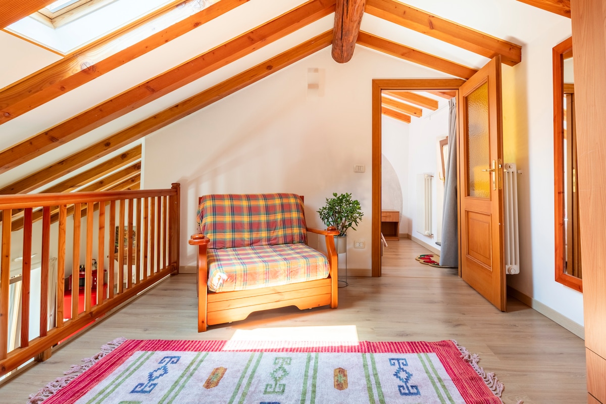 An inviting loft space features a wooden beamed ceiling and large skylights allowing natural light to brighten the area. A small checkered couch is placed on a patterned rug, providing a comfortable seating option. A wooden railing adds a warm touch to the open layout.