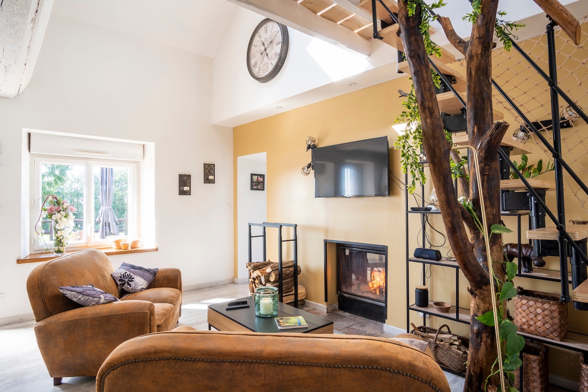 The living area features a warm color palette with a large window allowing natural light to fill the space. A modern fireplace is centered against a wall, flanked by a TV. Two comfortable chairs and a coffee table complement the open layout, accented by a staircase leading to the upper level.