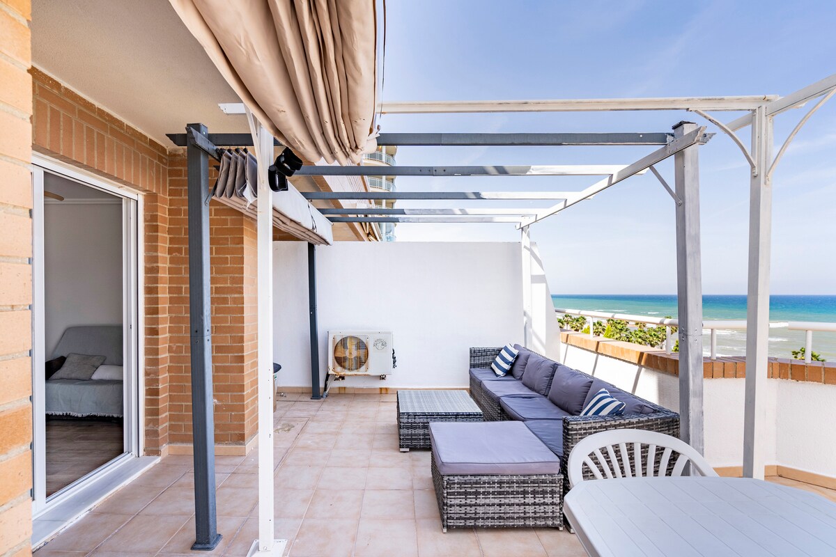 A spacious terrace features a shaded seating area with a large sectional sofa and a glass table. An unobstructed view of the ocean is visible, complemented by the clear blue sky. A small air conditioning unit is situated on the wall, adding convenience to the outdoor space.