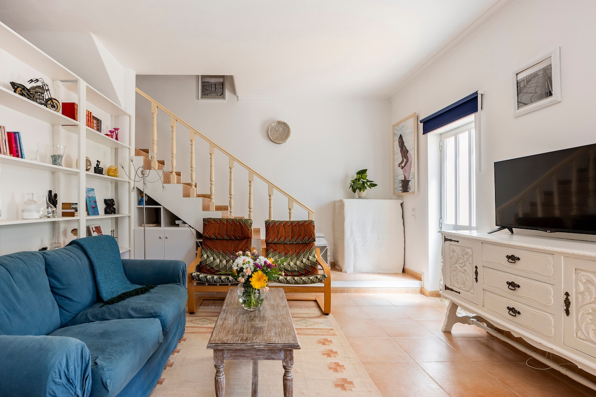 A bright living area displays a comfortable blue sofa and two patterned chairs. A wooden coffee table is centered on a neutral rug. A white shelving unit and a television unit enhance the space, while natural light enters through a window beside the staircase.