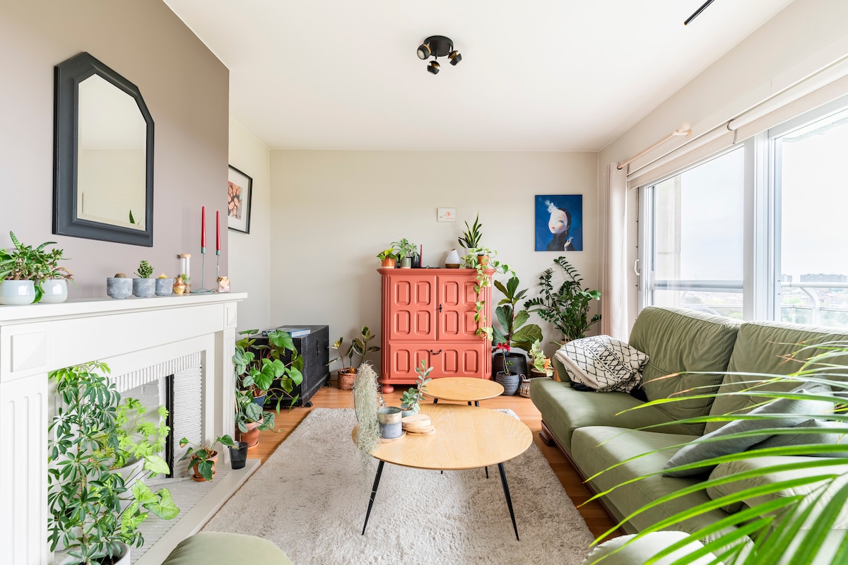 A bright living room features a green sofa accompanied by two round wooden coffee tables. A colorful cabinet stands against the wall, adorned with plants. Natural light floods the space through the large windows, creating a fresh and lively atmosphere among the diverse greenery.