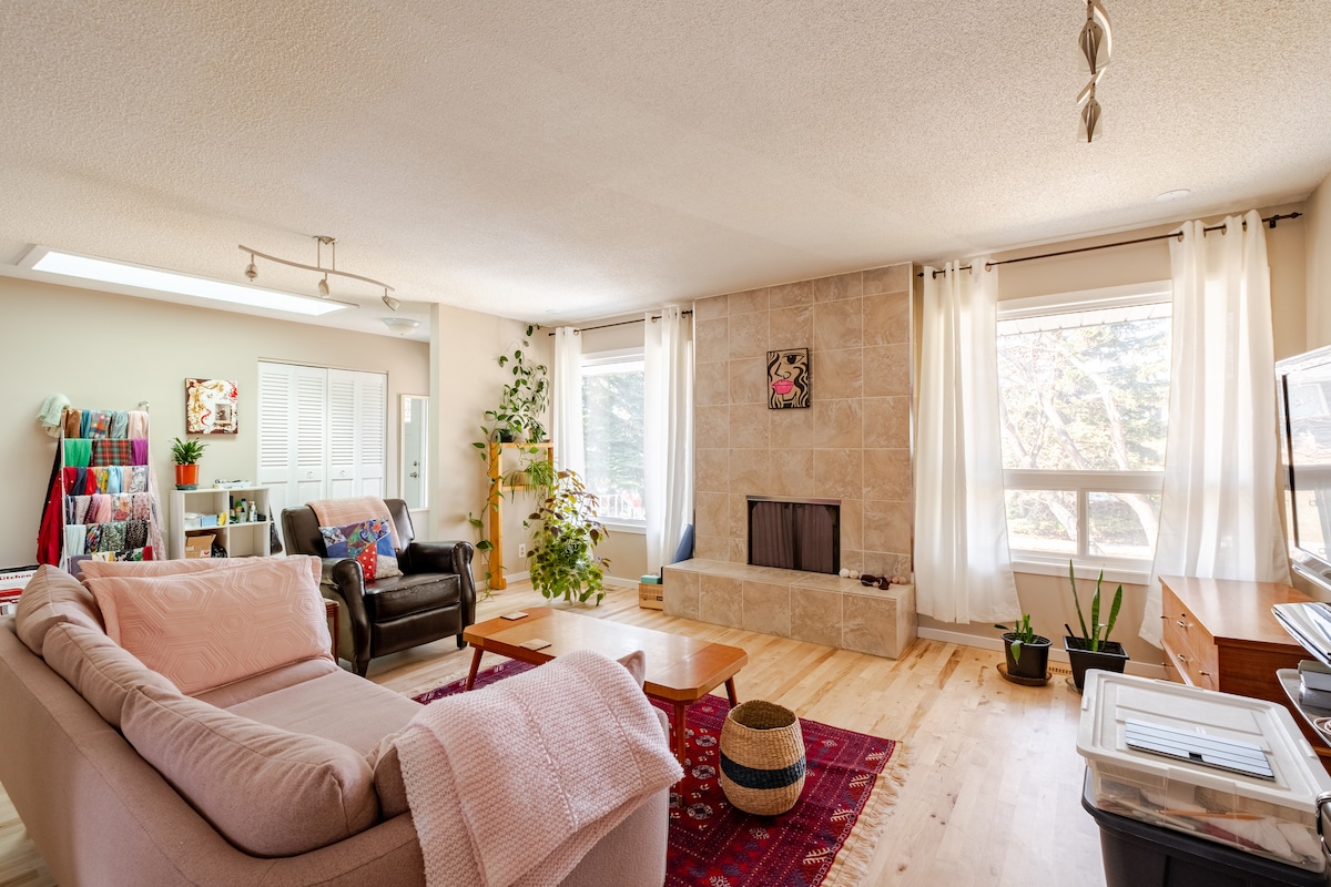 A spacious living area is highlighted by a stone fireplace, surrounded by large windows that bring in natural light. Comfortable seating options include a couch and armchair. Decorative elements such as houseplants and a patterned rug add a welcoming touch to the room.