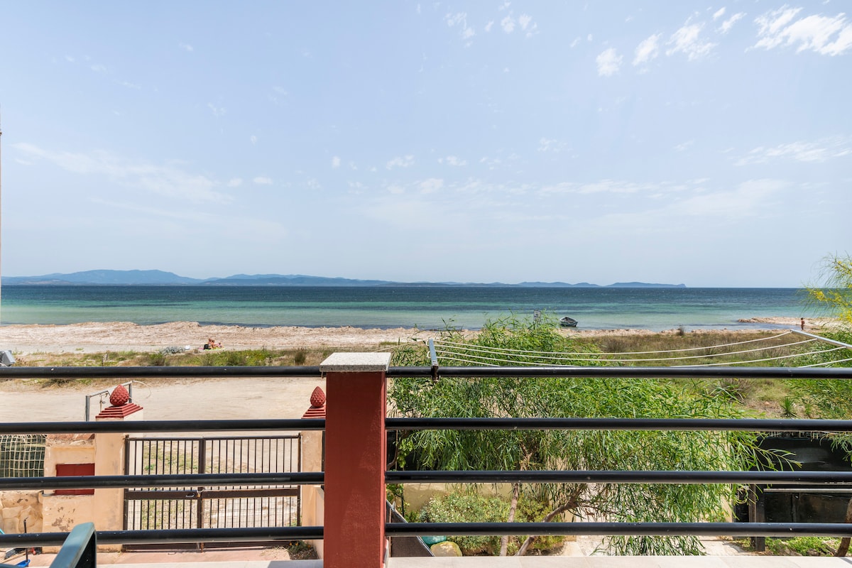 A view of the sea from a balcony, showcasing a tranquil shoreline and distant islands on the horizon. The water reflects shades of blue under a clear sky, while green foliage frames the scene. A sandy beach stretches along the coast, illustrating the peaceful environment.