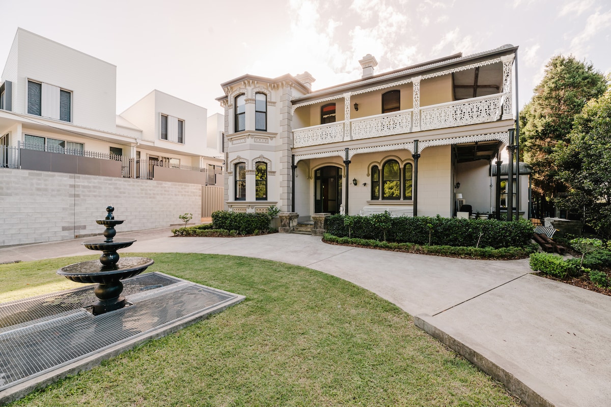 The grand mansion features a classic architecture with detailed balconies and ample greenery surrounding the property. A decorative fountain is positioned in the foreground, with a spacious lawn providing an inviting entrance. Natural light enhances the exterior details, contributing to the historic charm.