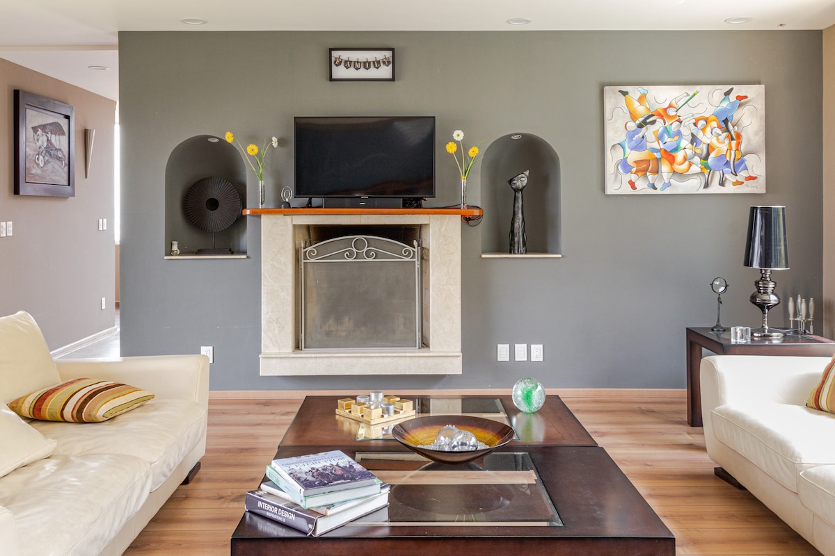 A modern living room features a sophisticated design with a light-colored sofa and a sleek coffee table adorned with decorative items. Above the fireplace, a television is mounted, complemented by artistic wall decor and stylish accents in built-in niches.