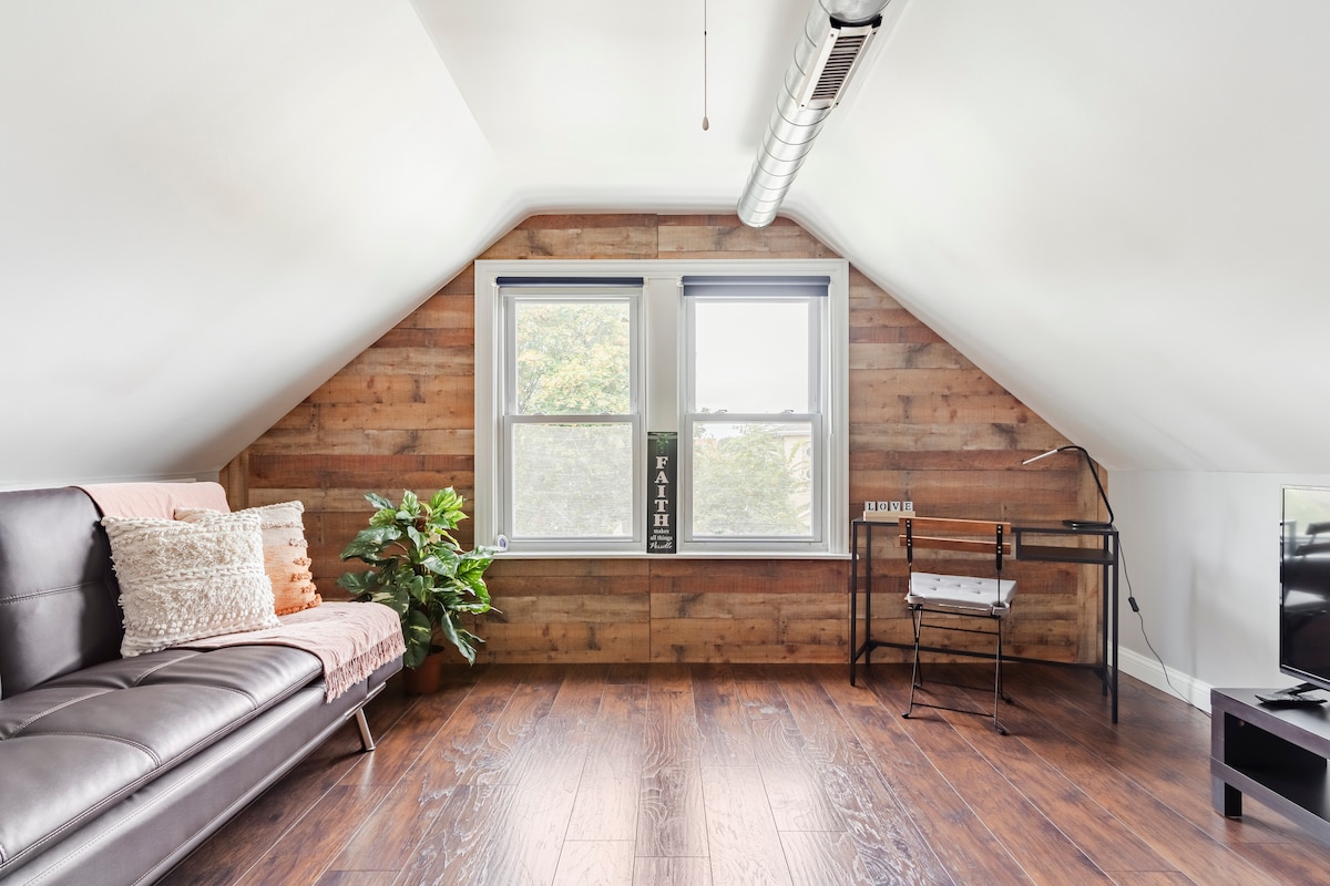 A bright and airy loft space is characterized by sloped ceilings and wooden paneling. A dark sofa is complemented by decorative pillows, while a small desk and chair are positioned by the window. Lush greenery adds a touch of nature to the cozy setting.