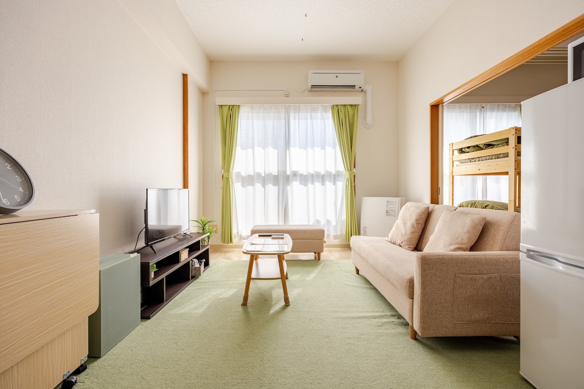 A well-lit living area features a light-colored sofa and a wooden table positioned on a soft green carpet. Large windows with sheer curtains allow natural light to fill the space. A TV is placed on a low cabinet, and a refrigerator is visible in the background.