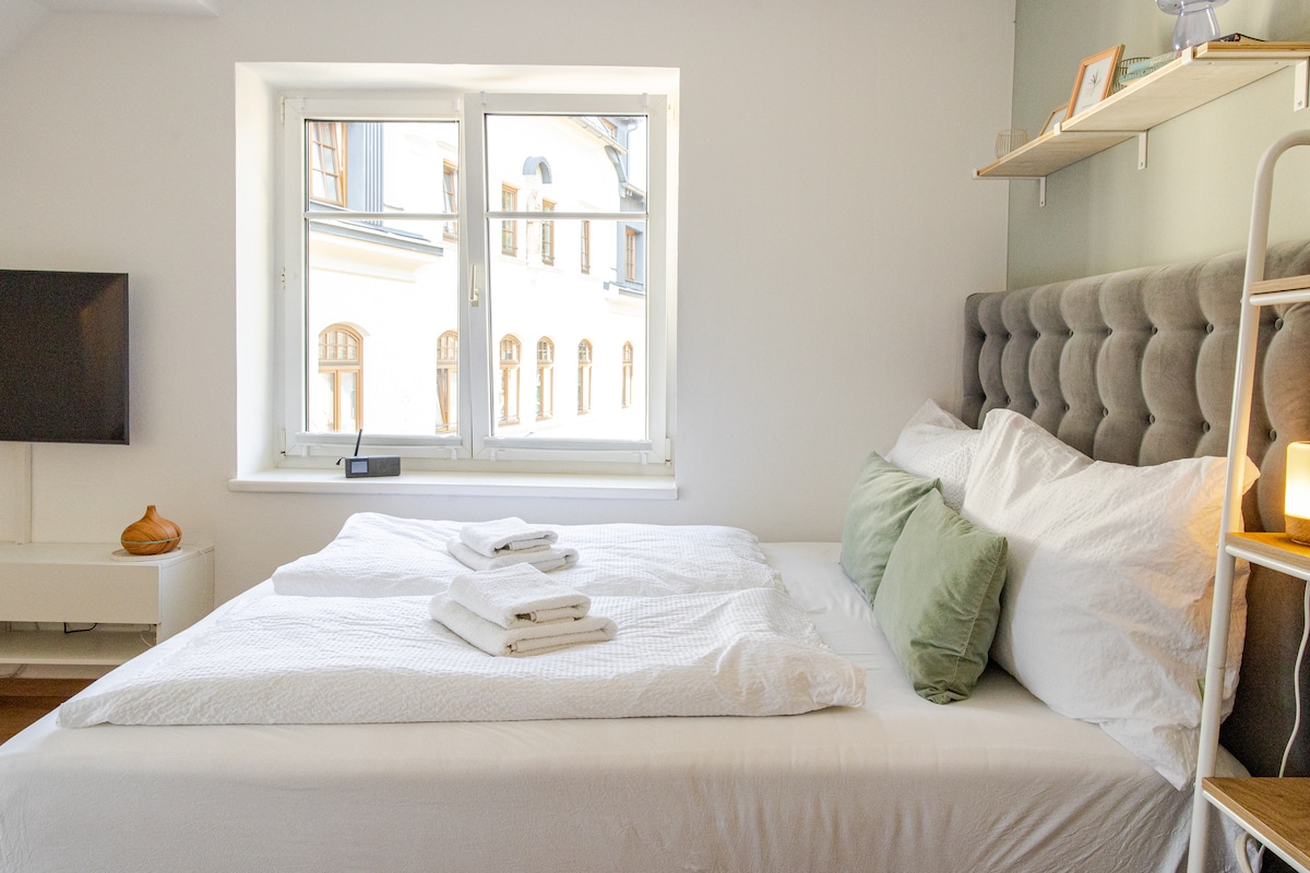 A bright sleeping area features a spacious bed dressed in light linens, with neatly arranged towels at the foot. Soft green pillows and a tufted headboard add a gentle touch. Natural light filters through a large window, illuminating the room's simple design.