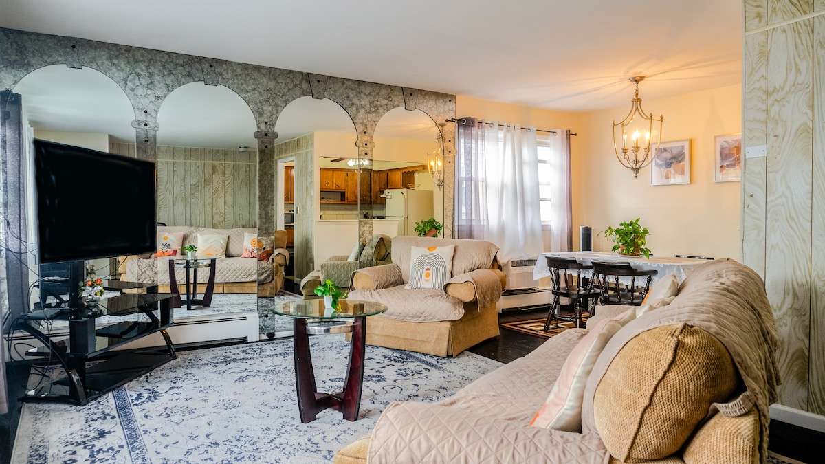 The living room features a spacious arrangement of seating with a large sofa and a glass coffee table centered on a textured area rug. Mirrored arches are present in the background, reflecting light and adding depth to the room. Natural light enters through sheer curtains.