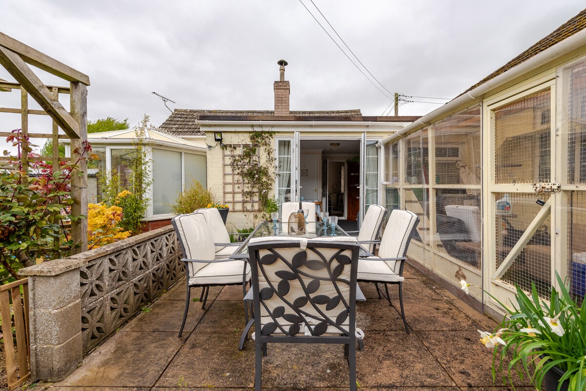An outdoor seating area is presented with a glass-top table surrounded by six cushioned chairs. The patio features a stone floor, and nearby, a trellis is adorned with greenery. A view into the house reveals bright interiors through open glass doors.