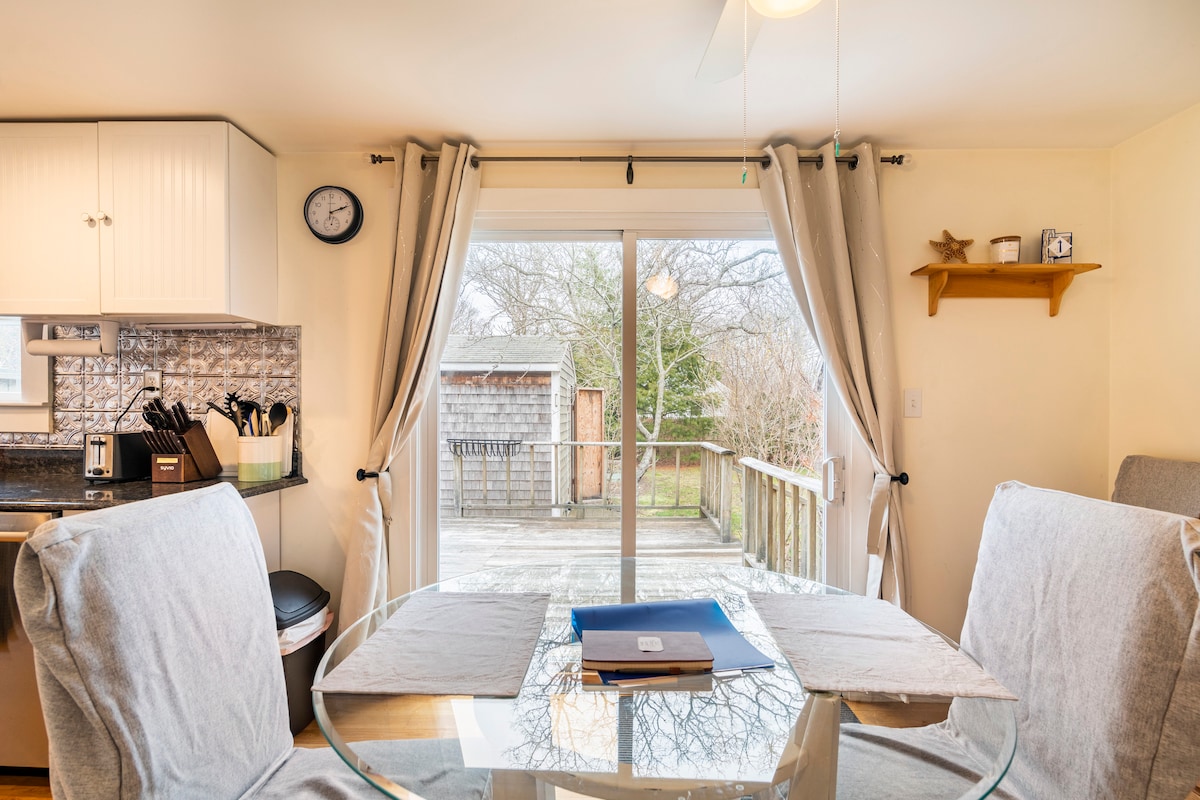 A dining area features a round glass table with light grey upholstered chairs. Natural light shines through the sliding glass door, which leads to a wooden deck and backyard. Minimalistic decor includes a clock on the wall and a small shelf with decorative items.