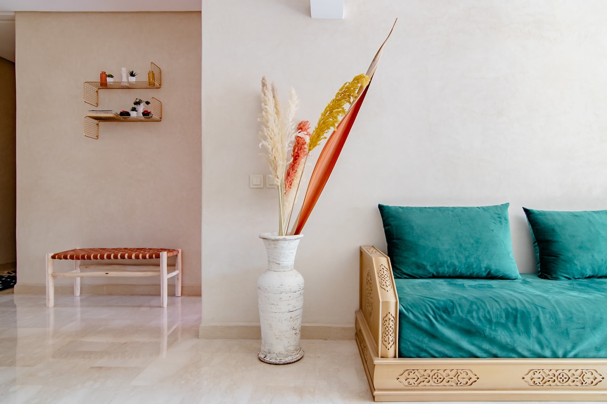 A seating area features a wooden couch with teal cushions, accompanied by a decorative vase holding tall, colorful pampas grass. A minimalist shelf with small plants is mounted on the wall nearby, while a bench with a patterned seat is placed against the opposite wall.