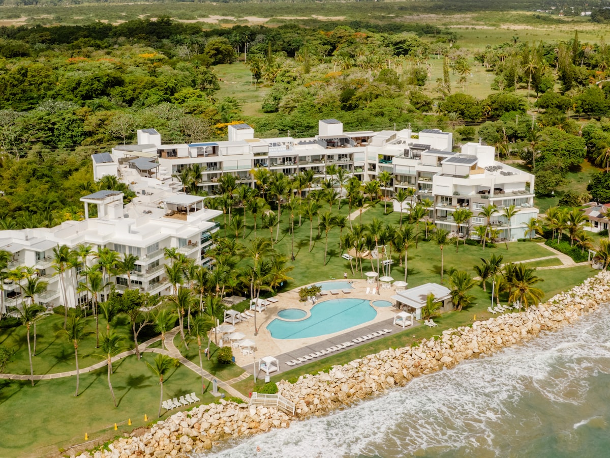 An aerial view captures the expansive Seawinds complex, showcasing modern white buildings surrounded by lush greenery and palm trees. A centrally located pool is visible amidst spacious grassy grounds, with lounge chairs arranged nearby, all overlooking the ocean and rocky shoreline.