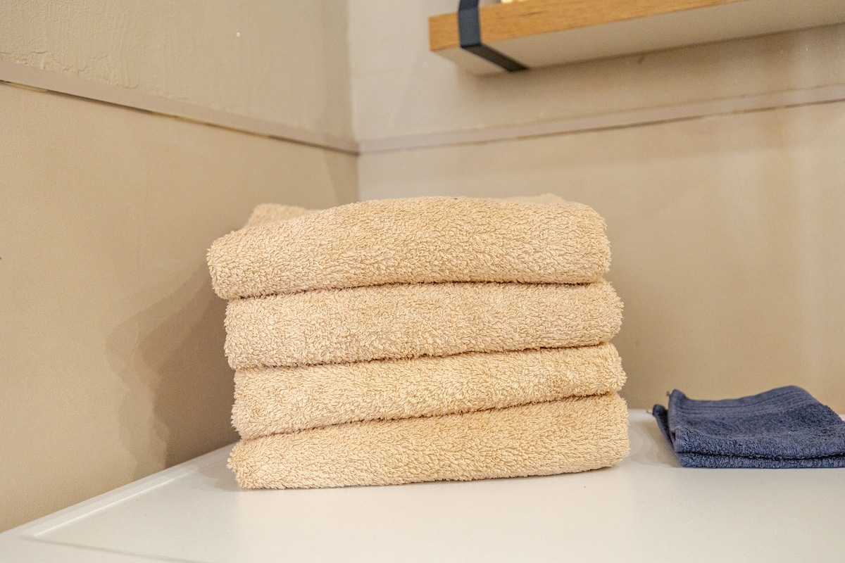 A neatly arranged stack of soft, beige towels is displayed on a white surface. Beside the stack, a single navy towel is positioned, providing a subtle contrast in color and texture.